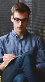 A young man in glasses writes in a notebook while sitting on a stylish couch indoors.