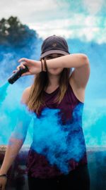 A stylish woman surrounded by bright blue smoke outdoors, creating a dynamic fashion statement.